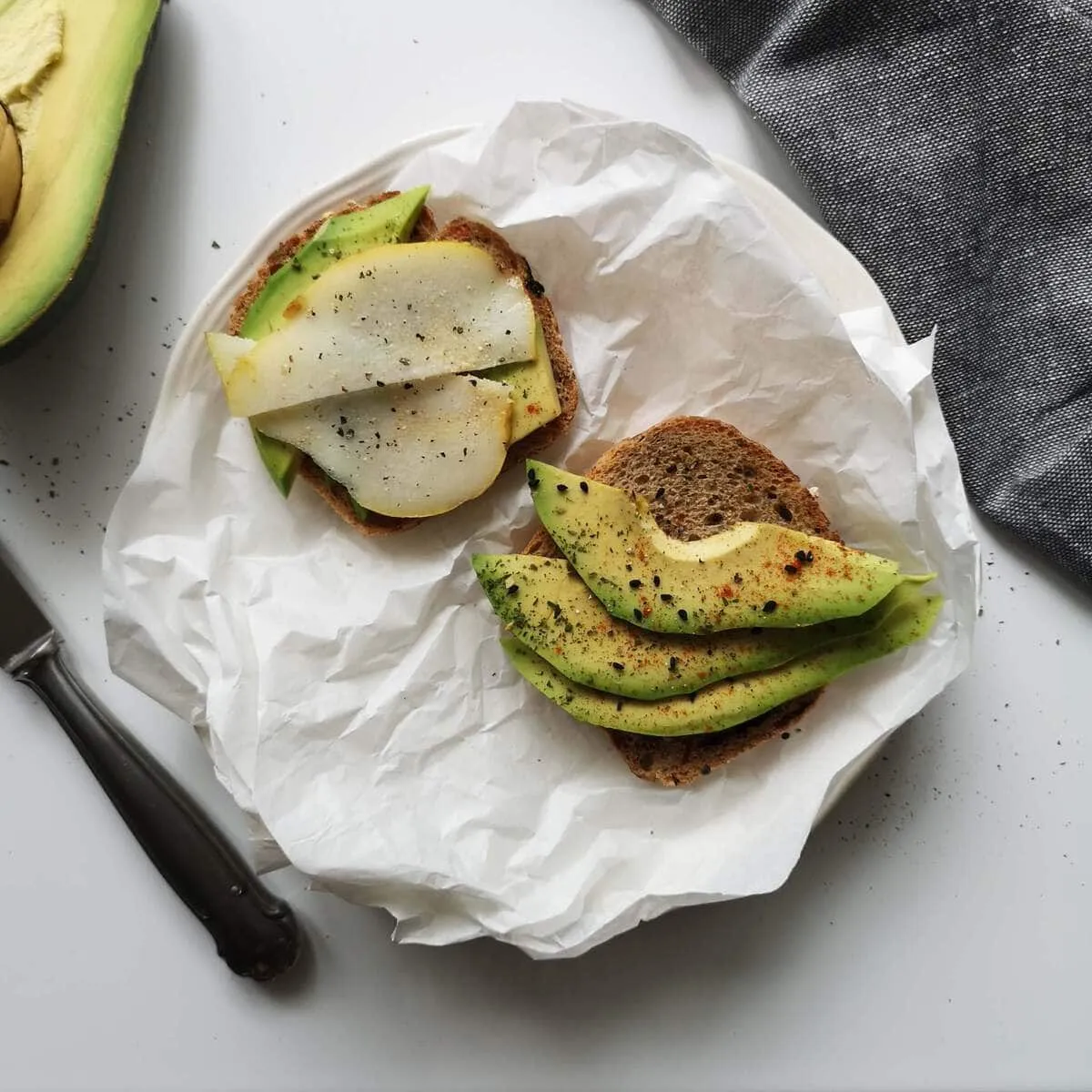 Deux tartines garnies d’avocat et de poire sur du papier, avec un couteau