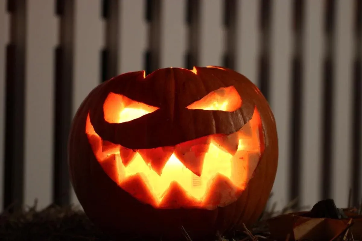 A lit jack-o’-lantern with a jagged carved grin before a white picket fence