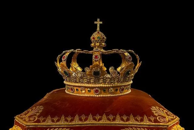 A jewelled gold crown resting on a red velvet cushion against a black background